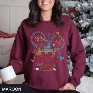 A woman wearing a maroon sweater with a Disney-themed design, including Mickey Mouse and fireworks, is smiling and holding a mug.