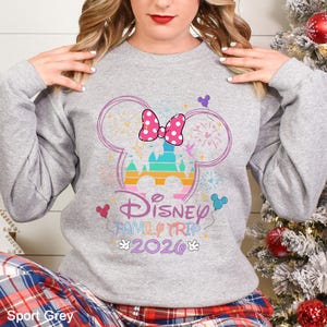 A woman wearing a gray sweatshirt with a Disney-themed design, including a Mickey Mouse ear and fireworks, posing in front of a Christmas tree.
