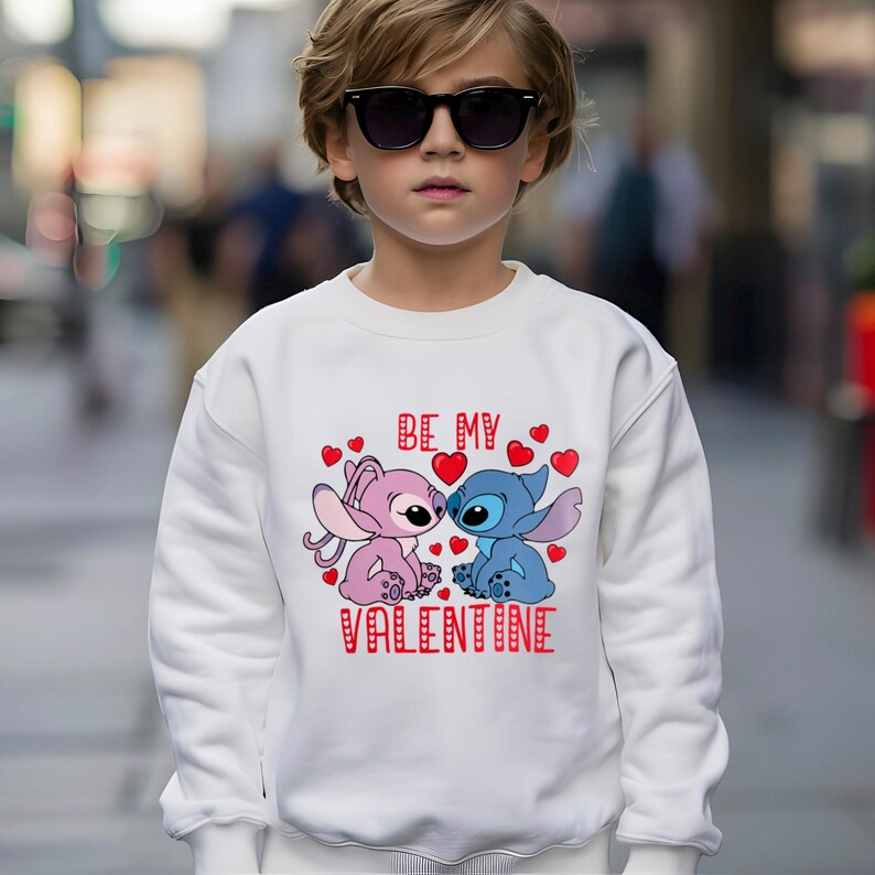 A young boy wearing a white sweatshirt with a graphic design featuring two cartoon characters, one pink and one blue, and the text "Be my Valentine".