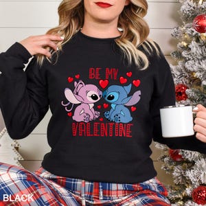 A woman wearing a black sweatshirt with a graphic design featuring two cartoon characters, one pink and one blue, along with the text "Be my Valentine".