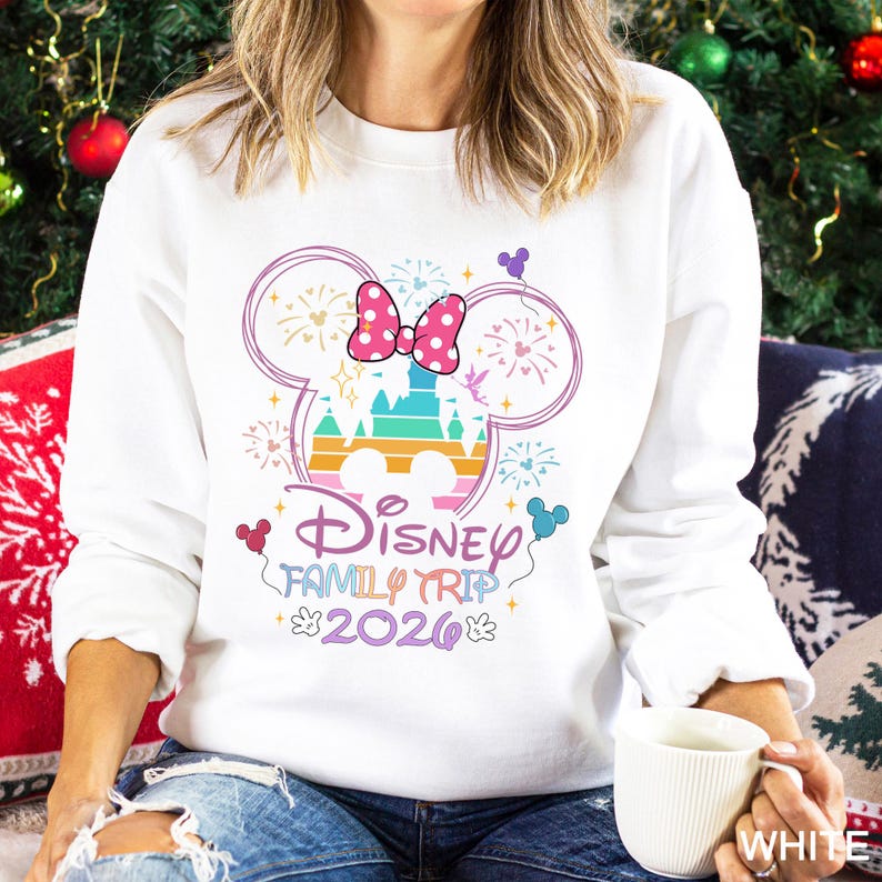 A woman wearing a white sweater with a Disney-themed design, including a Mickey Mouse ear and fireworks, is sitting on a couch with a mug in her hand.
