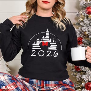 A woman wearing a black sweater with a white castle graphic and the year "2026" printed on it, holding a white mug.