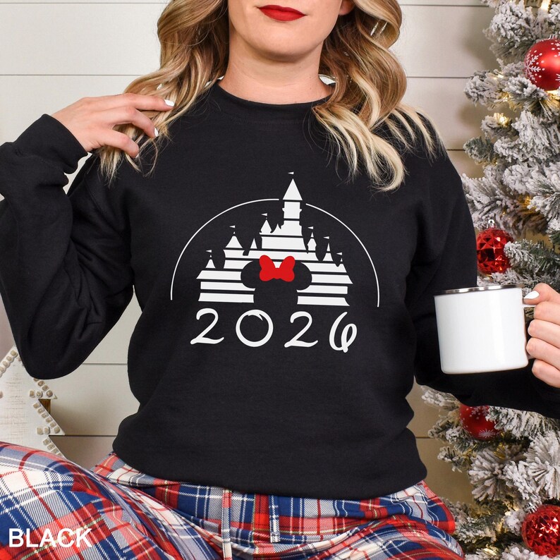 A woman wearing a black sweater with a white castle graphic and the year "2026" printed on it, holding a white mug.