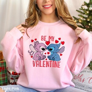 A woman wearing a pink sweatshirt with a graphic of two cartoon characters, one purple and one blue, and the text "Be My Valentine" printed on it.