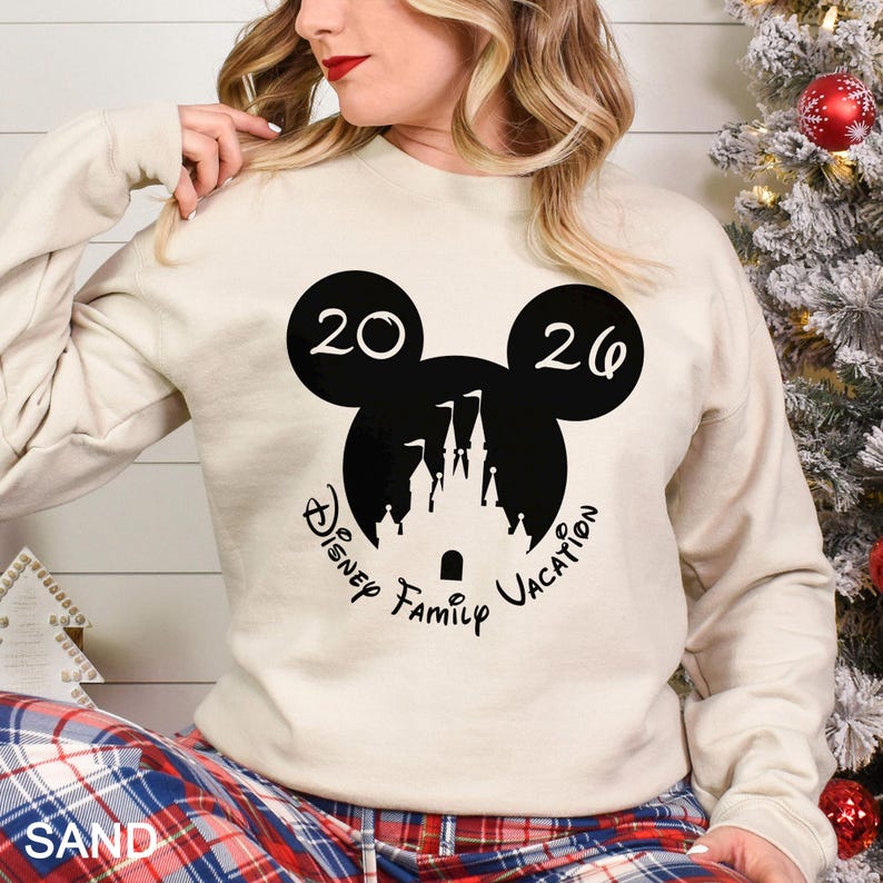 A woman wearing a white sweatshirt with a black Mickey Mouse logo and text, sitting next to a Christmas tree.