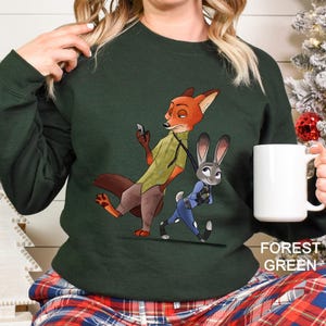 A woman wearing a green sweater with an illustration of a fox and a rabbit on it, holding a white mug.
