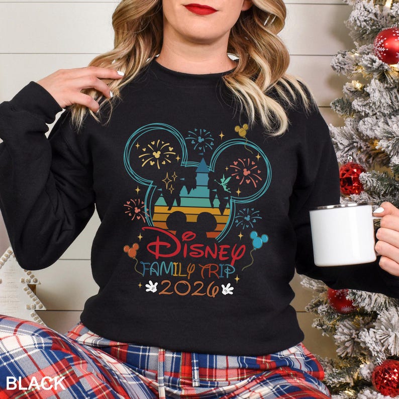 A woman wearing a black sweater with a Disney-themed design and text, holding a white mug, with a Christmas tree in the background.