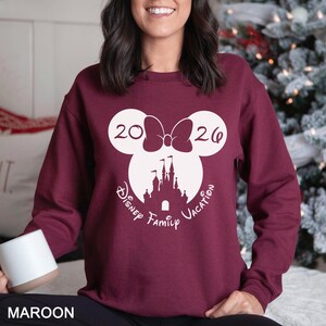 A woman wearing a burgundy sweater with a white Mickey Mouse design and text that says "2026 Disney Family Vacation".