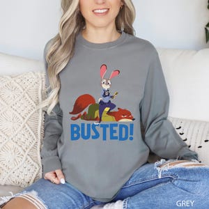 A woman wearing a gray long-sleeved shirt with a cartoon character and the text "BUSTED!" printed on it.