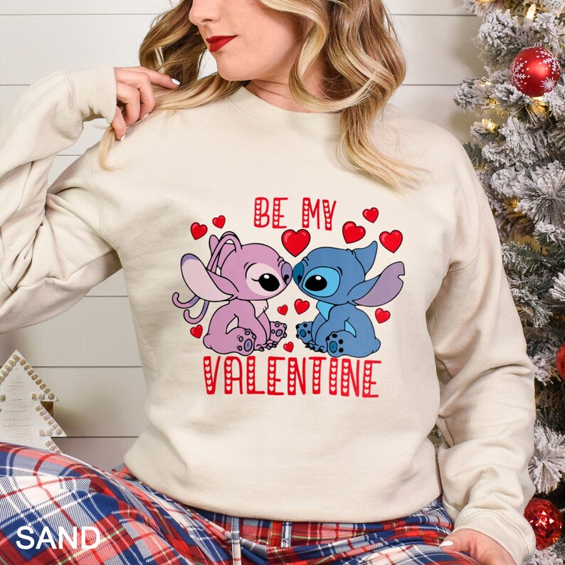 A woman wearing a white sweatshirt with a graphic design featuring two cartoon characters, one pink and one blue, along with the text "Be my Valentine".