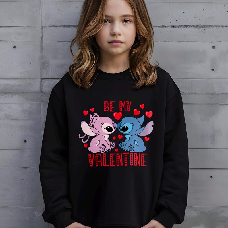 A young girl wearing a black sweatshirt with a graphic design featuring two cartoon characters, one pink and one blue, along with the text "Be my Valentine".