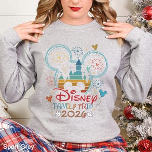 A woman wearing a gray sweatshirt with a Disney-themed design, including a Mickey Mouse and fireworks, posing in front of a Christmas tree.