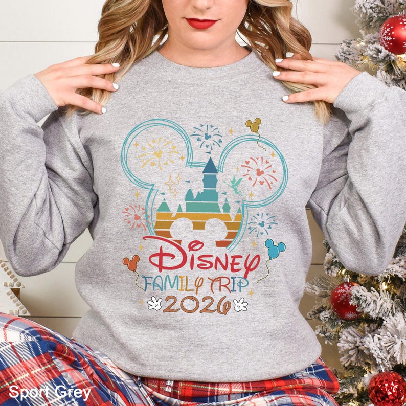 A woman wearing a gray sweatshirt with a Disney-themed design, including a Mickey Mouse and fireworks, posing in front of a Christmas tree.