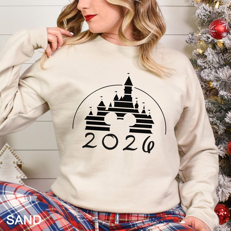 A woman wearing a white sweatshirt with a black graphic of a castle and the year "2020" printed on it, standing next to a Christmas tree.