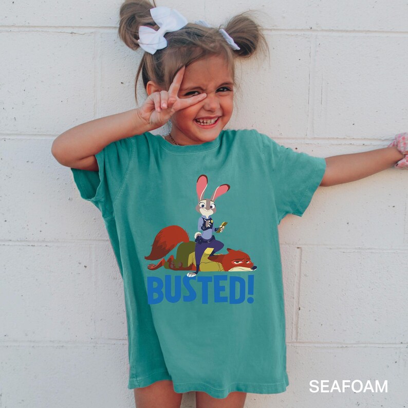 A young girl wearing a green t-shirt with a cartoon rabbit character and the text "BUSTED!" is smiling and making a peace sign.