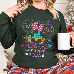 A woman wearing a green sweater with a Disney-themed design, including a Mickey Mouse ear and fireworks, holding a white mug.
