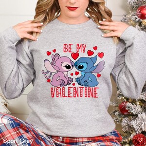 A woman wearing a gray sweatshirt with a graphic design featuring two cartoon characters, a pink one and a blue one, along with the text "Be My Valentine".