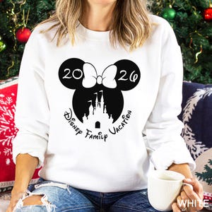 A woman wearing a white sweater with a graphic design of Minnie Mouse and a castle, along with the text "Disney Family Vacation" printed on it. She is sitting on a couch with Christmas decorations visible in the background.