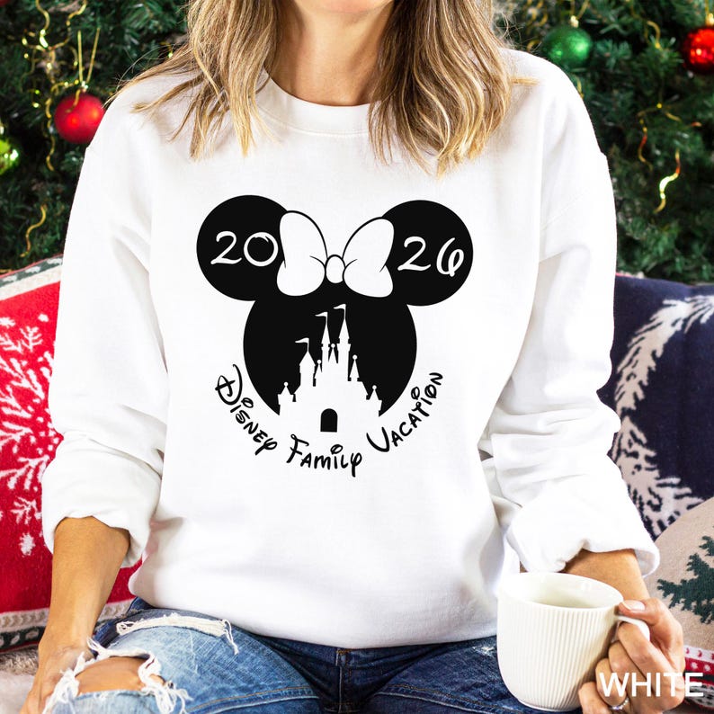 A woman wearing a white sweater with a graphic design of Minnie Mouse and a castle, along with the text "Disney Family Vacation" printed on it. She is sitting on a couch with Christmas decorations visible in the background.