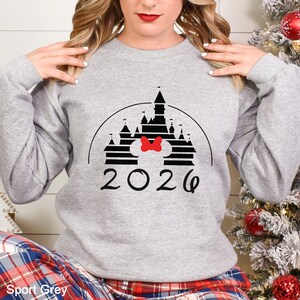 A woman wearing a grey sweatshirt with a graphic of a castle and the year "2026" printed on it.
