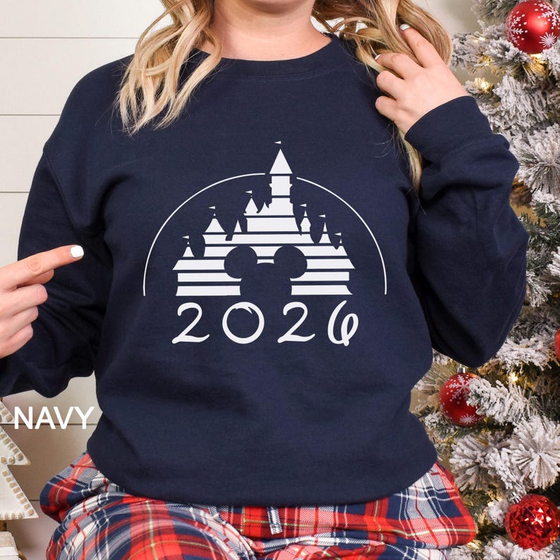 A woman wearing a navy blue sweatshirt with a white graphic of a castle and the year "2026" printed on it, standing in front of a Christmas tree.