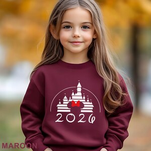 A young girl with long blonde hair wearing a maroon sweater with a white castle graphic and the year "2026" printed on it.