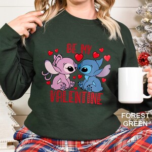 A woman wearing a green sweatshirt with a graphic of two cartoon characters, one pink and one blue, and the text "Be My Valentine" printed on it. She is holding a white mug.