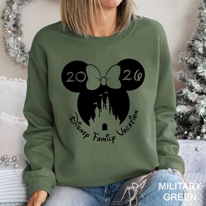 A woman wearing a green sweatshirt with a graphic design featuring a Minnie Mouse and a castle, along with the text "Disney Family Vacation".