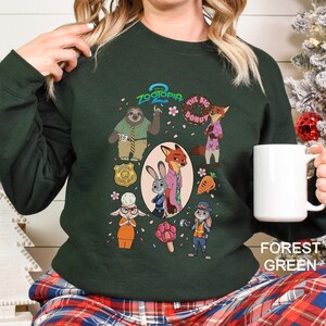 A woman wearing a green sweatshirt with a graphic of various cartoon characters from the movie "Zoografia". She is holding a white mug in her hand.