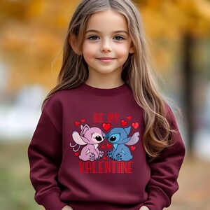 A young girl wearing a burgundy sweater with a graphic of two cartoon characters, one pink and one blue, and the text "Be my Valentine" printed on it.