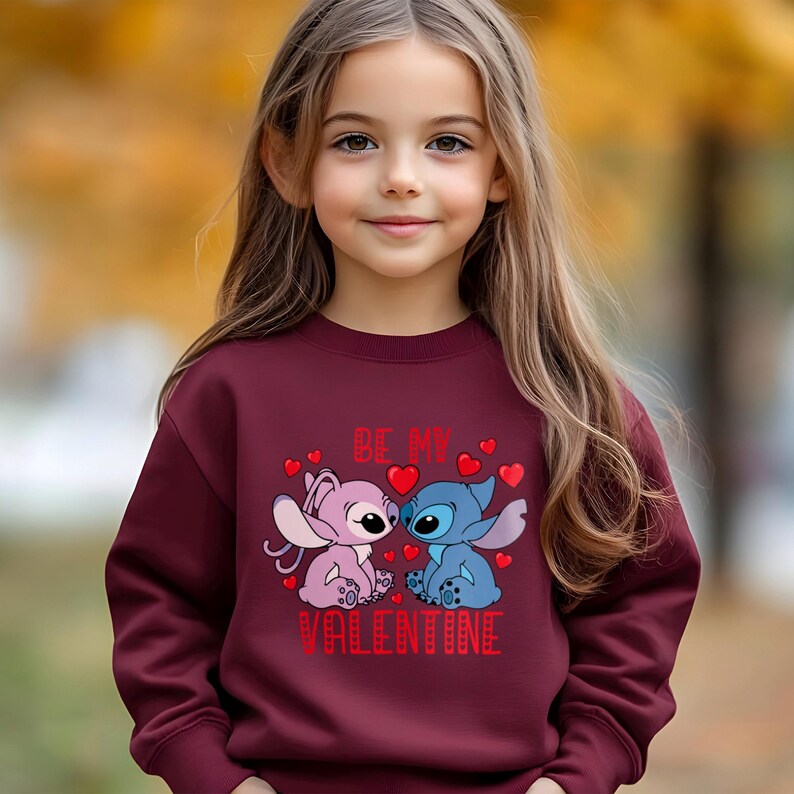A young girl wearing a burgundy sweater with a graphic of two cartoon characters, one pink and one blue, and the text "Be my Valentine" printed on it.