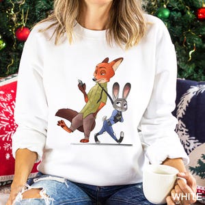 A woman wearing a white sweatshirt with an illustration of a fox and a rabbit on it, holding a white mug.