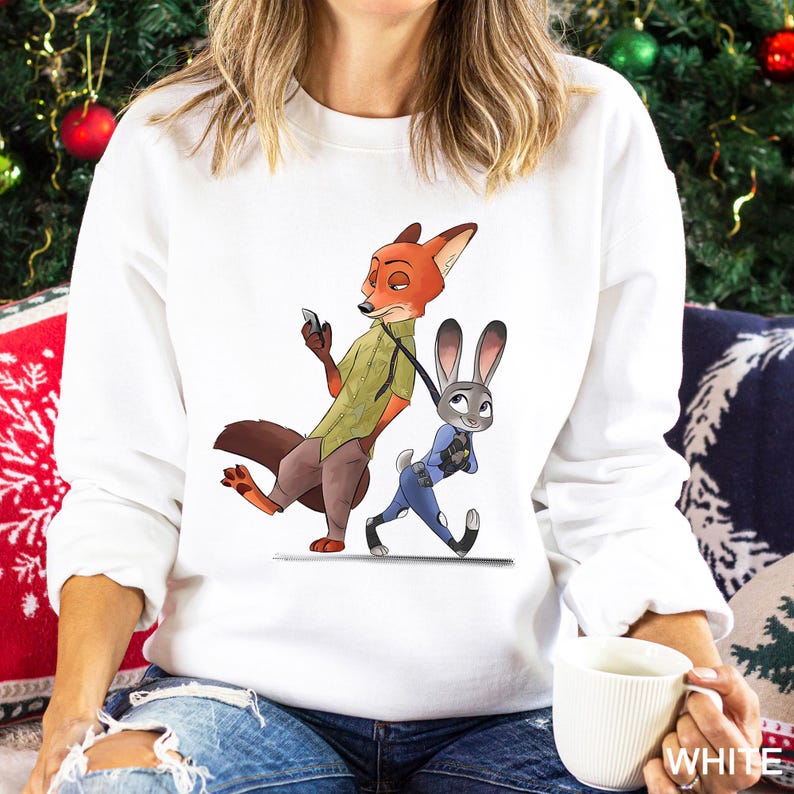 A woman wearing a white sweatshirt with an illustration of a fox and a rabbit on it, holding a white mug.