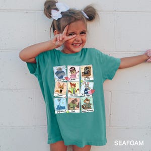 A young girl wearing a green t-shirt with a graphic of cartoon characters and text, smiling and making a peace sign gesture.