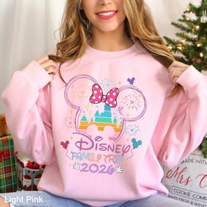 A woman wearing a pink sweatshirt with a Disney-themed design, including a Mickey Mouse ear and fireworks, smiling and posing in front of a Christmas tree.