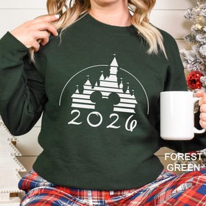 A woman wearing a green sweater with a white graphic of a castle and the year "2026" printed on it, holding a white mug.
