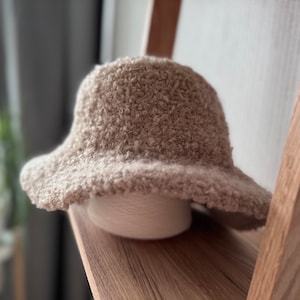 May include: A beige, textured bucket hat with a wide brim, resting on a white spool of yarn. The hat has a soft, fuzzy appearance. The hat is displayed on a wooden shelf.