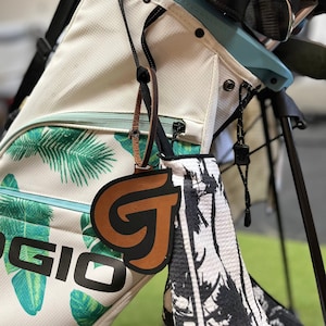 Könnte beinhalten: Eine weiße Golftasche mit tropischem Blatt-Muster und einem schwarzen "OGIO"-Logo. Ein brauner Leder-Golfball-Marker mit einem "G"-Logo ist an der Tasche befestigt. Die Tasche hat einen blau-weißen Gurt und einen blauen Plastikhaken.