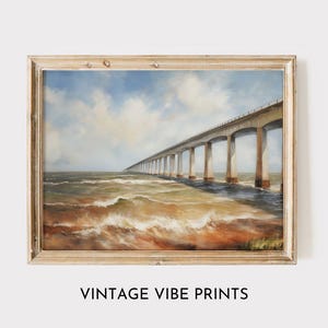 Op de afbeelding: Een ingelijste vintage-stijl print die een lange brug over een golvende oceaan onder een bewolkte hemel afbeeldt. Het kunstwerk zit in een houten lijst. De tekst "VINTAGE VIBE PRINTS" staat onderaan de afbeelding.