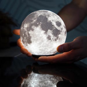 Może przedstawiać: Sferyczna, podświetlana lampa księżycowa o szczegółowej powierzchni, trzymana w dłoniach. Widoczne są kratery i tekstury lampy, tworząc realistyczny wygląd księżyca. Lampa odbija się na ciemnej powierzchni.