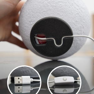 May include: A white, spherical lamp with a textured surface. The lamp has a black base with a white cord and a USB connector. The cord also features an on/off switch. The lamp is designed to resemble the moon.