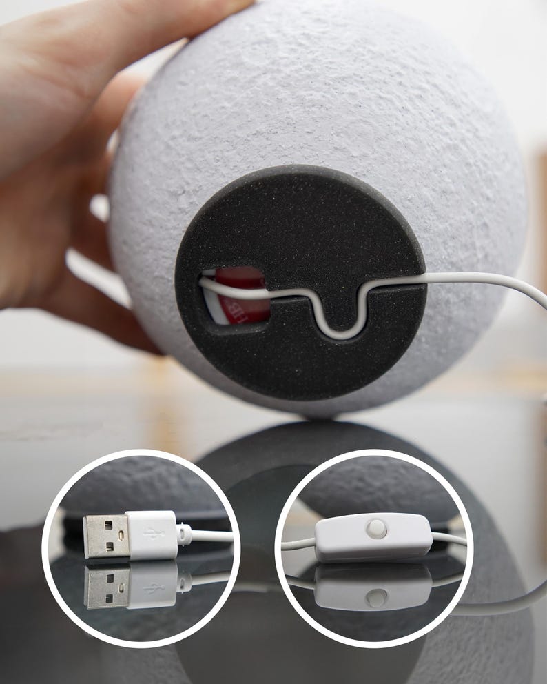 May include: A white, spherical lamp with a textured surface. The lamp has a black base with a white cord and a USB connector. The cord also features an on/off switch. The lamp is designed to resemble the moon.