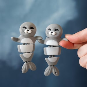 Peut inclure: Deux figurines de phoque grises et blanches avec des corps articulés et des visages souriants. Les phoques ont les bras tendus et un design segmenté, avec une nageoire caudale. Le fond est un dégradé de bleu.
