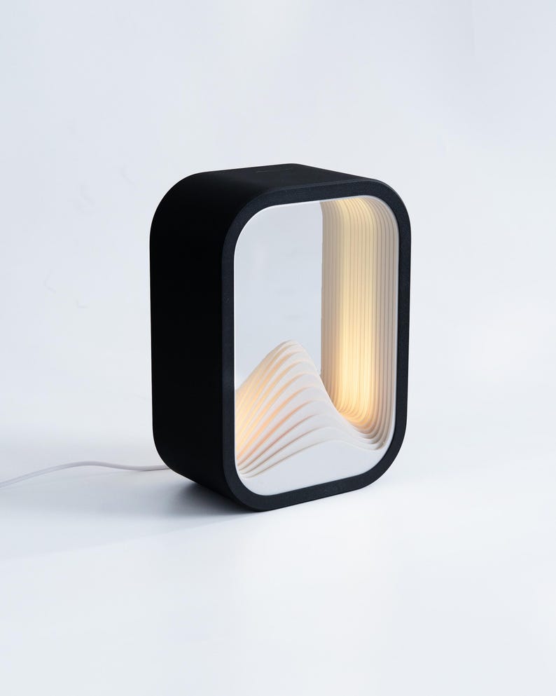 May include: A rectangular table lamp with a matte black outer frame and a white interior featuring layered, wave-like ridges. The lamp emits a warm, soft glow from within the structure, which is connected to a thin white power cord.