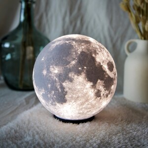 May include: A spherical moon lamp with a detailed surface texture, emitting a soft, warm glow. The lamp sits on a black base, creating a celestial ambiance. Decorative vases are in the background.