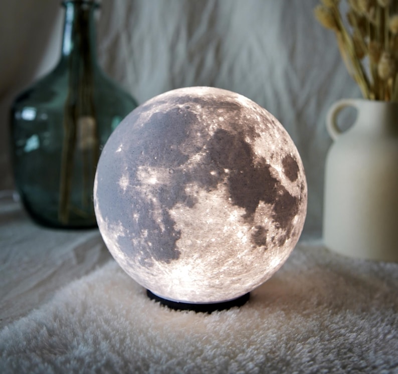 May include: A spherical moon lamp with a detailed surface texture, emitting a soft, warm glow. The lamp sits on a black base, creating a celestial ambiance. Decorative vases are in the background.
