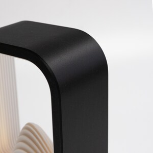 May include: A black, rectangular frame with rounded corners, featuring a smooth, matte finish. Inside the frame, there are multiple, thin, white, wavy, layered panels that create a textured, undulating appearance within the structure.