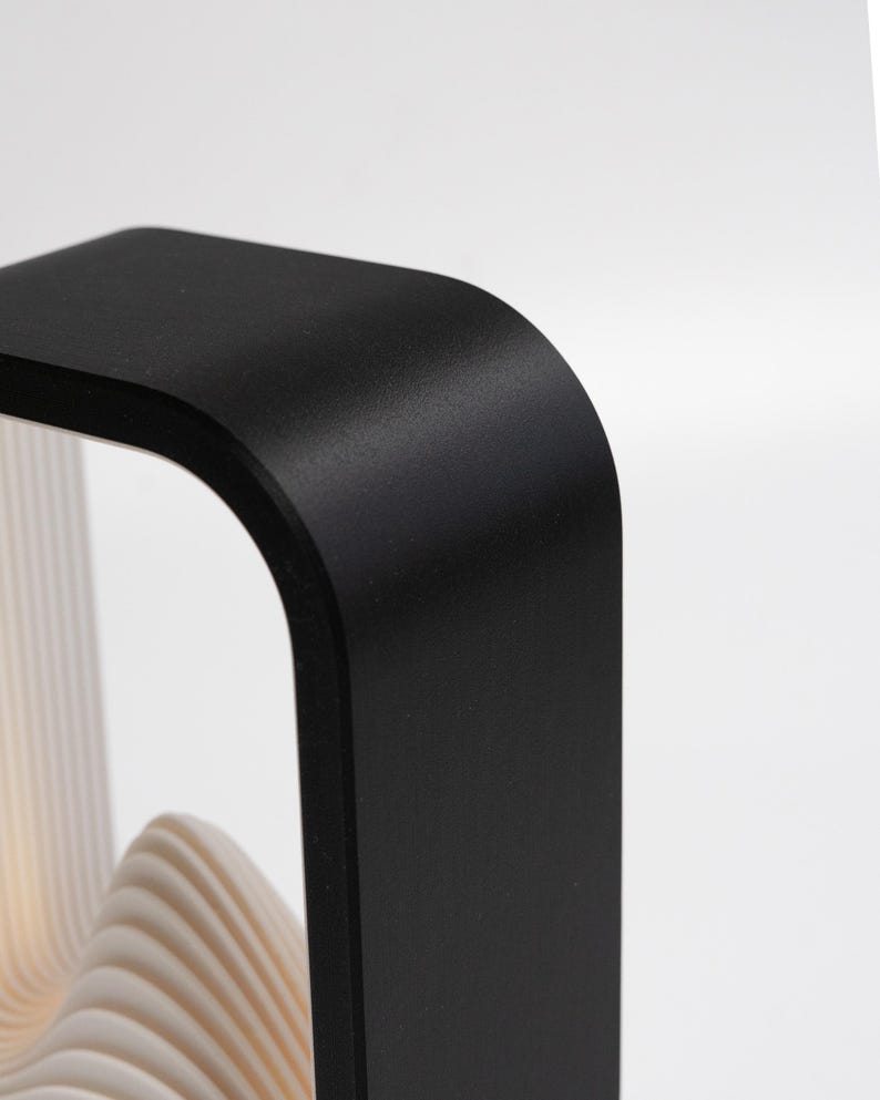 May include: A black, rectangular frame with rounded corners, featuring a smooth, matte finish. Inside the frame, there are multiple, thin, white, wavy, layered panels that create a textured, undulating appearance within the structure.