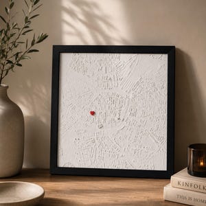 Puede incluir: Una pieza de arte mural cuadrada con marco negro que presenta un mapa en relieve tridimensional blanco de un trazado urbano. Un pequeño alfiler rojo brillante en forma de corazón está fijado en el centro de la superficie blanca texturizada.