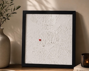 May include: A square, black-framed wall art piece featuring a white, three-dimensional relief map of a city street layout. A small, bright red heart-shaped pin is attached to the center of the textured white map surface.